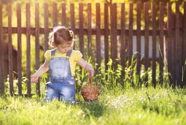A little girl sitting in the grass with a wood fence behind her.